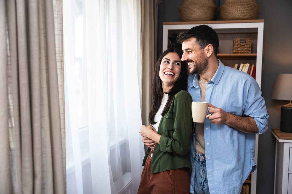 Young couple standing near window at their new apartment, enjoying their new life together. Engaged young people man and woman taking time for them to relax
