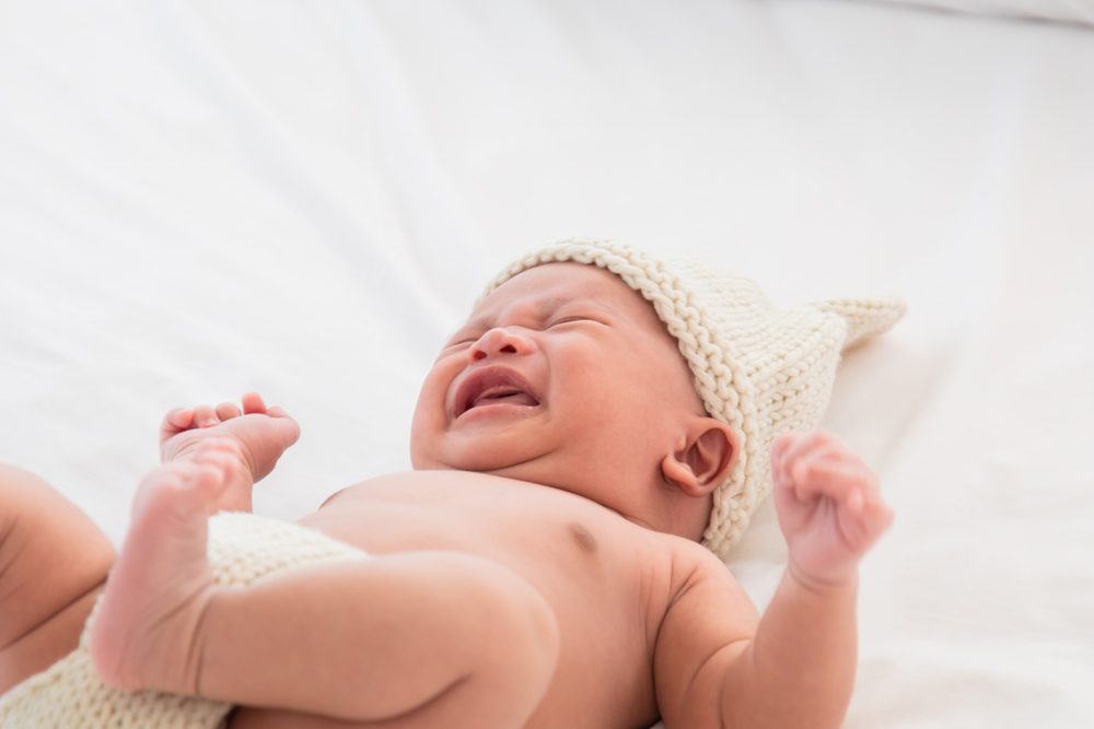 Newborn baby crying lying on white bed. Adorable infant 0-1month fussy screaming unhappy angry. Child wearing pants and rabbit hat beige knitted tired and hungry.