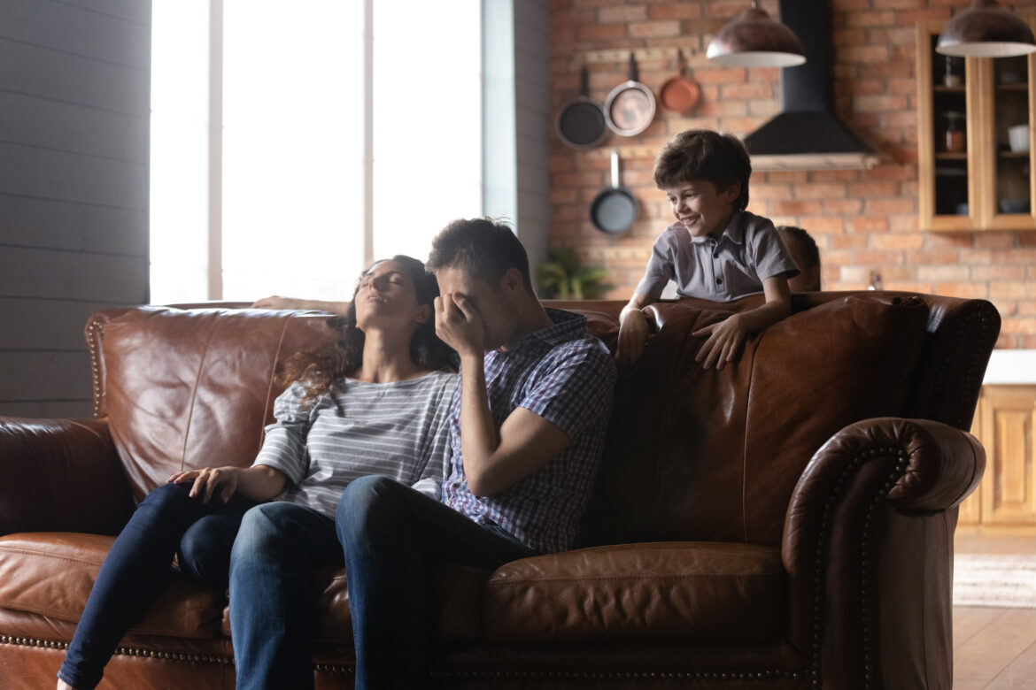 upset couple sitting on the couch and their kid behind them representing the concept of parental burnout