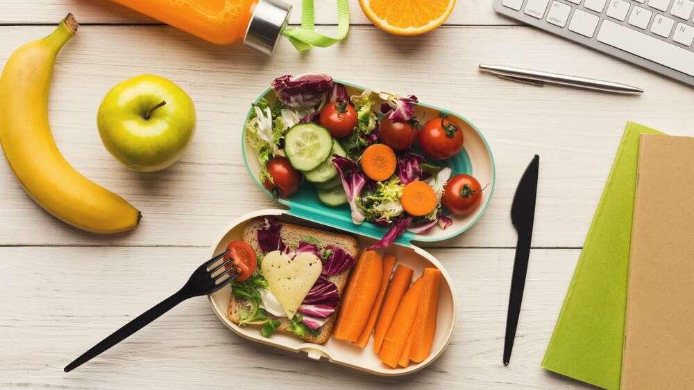 A tiffin  ox full of healthy vegetables and food surrounded by fruits and work essentials